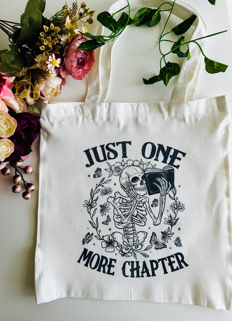 Pu&ograve; includere: Borsa di tela bianca con testo nero e un'immagine di uno scheletro che legge un libro. Il testo dice "Just One More Chapter".