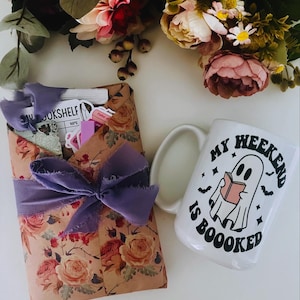 May include: A white ceramic mug with the text "MY WEEKEND IS BOOOOKED" and a ghost reading a book. A package wrapped in floral paper and tied with a purple ribbon sits beside the mug. Flowers are in the background.