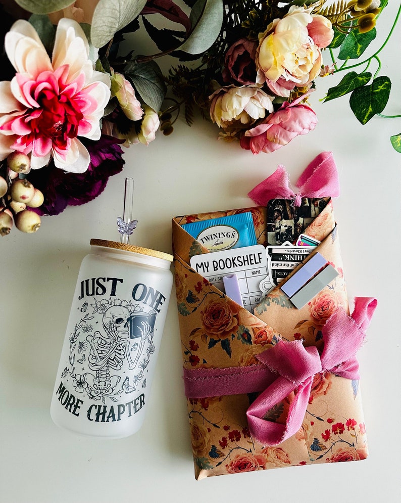 Pu&ograve; includere: Un regalo avvolto in carta marrone con un motivo floreale e un nastro rosa. Il regalo include un bicchiere di vetro trasparente con uno scheletro nero che legge un libro e il testo "Just One More Chapter". Il regalo include anche una scatola di t&egrave;, un registro dei libri e alcuni adesivi.