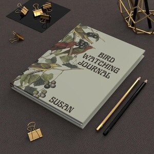 K&ouml;nnte beinhalten: Ein Hardcover-Vogelbeobachtungsjournal mit einer Cover-Illustration von V&ouml;geln, Bl&auml;ttern und Beeren. Das Journal tr&auml;gt den Titel "Bird Watching Journal" mit dem Namen "Susan" darunter. Goldene und schwarze Bleistifte und goldene B&uuml;roklammern sind ebenfalls sichtbar.