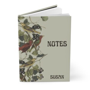 K&ouml;nnte beinhalten: Ein Notizbuch mit Hartcover und einem Design zum Thema Natur. Das Cover zeigt Illustrationen von V&ouml;geln, Trauben und Bl&auml;ttern in Gelb-, Rot- und Gr&uuml;nt&ouml;nen auf hellgr&uuml;nem Hintergrund. Das Wort "NOTES" und der Name "SUSAN" sind auf dem Cover aufgedruckt.