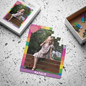 May include: A custom jigsaw puzzle featuring a photograph of a young child with blonde hair wearing a floral dress, sitting on a wooden fence. The puzzle border has a colorful striped pattern with a pink heart and the name NATALIE printed at the bottom.
