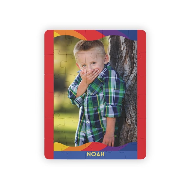 May include: A rectangular jigsaw puzzle featuring a photograph of a young child wearing a green and blue plaid button-down shirt, with hands covering the mouth. The puzzle has a colorful border and the name NOAH printed in yellow at the bottom.