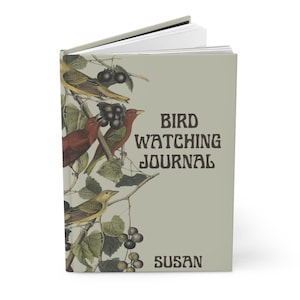 K&ouml;nnte beinhalten: Ein Hardcover-Vogelbeobachtungsjournal mit hellgr&uuml;nem Einband. Der Einband zeigt Illustrationen von bunten V&ouml;geln, Trauben und Bl&auml;ttern. Der Text "BIRD WATCHING JOURNAL" und der Name "SUSAN" sind auf dem Einband aufgedruckt.