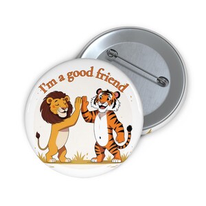 May include: A white button featuring a cartoon lion and tiger giving a high five. The lion is tan with a brown mane, and the tiger has orange and black stripes. Text reads "I'm a good friend."