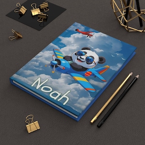 Puede incluir: Un diario de tapa dura azul con un panda de dibujos animados pilotando un avión azul contra un cielo nublado. El nombre "Noah" está impreso en blanco en la portada. Un biplano rojo vuela en el fondo.