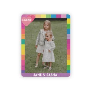 May include: A rectangular jigsaw puzzle featuring a photograph of two children standing on grass. The puzzle has a colorful border, a pink heart graphic with the text COUSINS, and the names JANE & SASHA printed at the bottom in white text.
