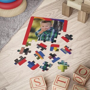 May include: A custom jigsaw puzzle featuring a photograph of a young child wearing a green and blue plaid shirt with their hands over their mouth. The puzzle is partially assembled with several loose pieces scattered below, one of which displays the name Noah.