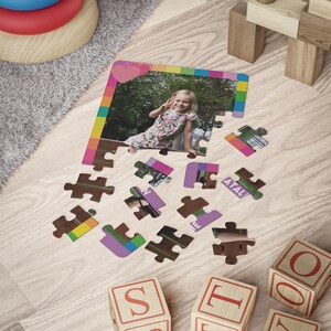 May include: A custom jigsaw puzzle featuring a photograph of a young child in a floral dress making a peace sign. The puzzle has a colorful border with a pink heart at the top and several detached puzzle pieces scattered below the main frame.