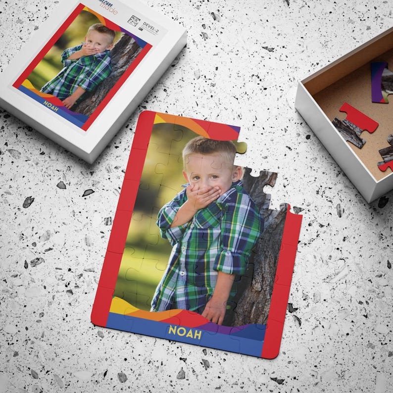 May include: A custom jigsaw puzzle featuring a photograph of a young child wearing a green and blue plaid shirt, with the name NOAH printed in yellow on a blue banner at the bottom. The puzzle is shown partially assembled with a matching box.