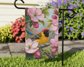 May include: A garden flag featuring a vibrant illustration of a yellow bird with a red head and blue tail perched on a branch. The bird is surrounded by pink blossoms and green leaves. The flag is displayed on a black metal stand in a garden setting.