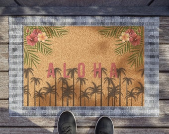 May include: A brown doormat with the word "ALOHA" in pink letters and a tropical leaf design. The doormat is made of coir and has a checkered pattern border.