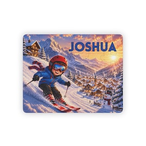 May include: A rectangular jigsaw puzzle featuring a cartoon skier in a blue jacket, red helmet, and goggles on a snowy slope. The name JOSHUA is printed in blue at the top. The background shows a mountain village, snow-covered trees, and a snowman.