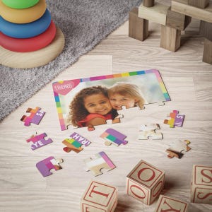May include: A rectangular jigsaw puzzle featuring a photograph of two children smiling, with the word FRIENDS printed inside a pink heart. The puzzle border has a colorful striped pattern, and several loose puzzle pieces are scattered nearby.