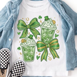 May include: White t-shirt with a St. Patrick's Day design featuring green bows, shamrocks, and green drinks with whipped cream. The shirt is paired with a light blue denim jacket and black and white checkered slip-on shoes.