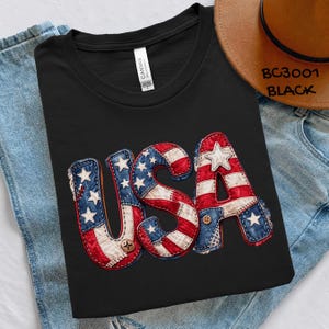 May include: Black t-shirt with the word "USA" in a patriotic design. The letters are made to look like they are sewn together with red, white, and blue fabric, and have button accents. A brown hat and blue jeans are in the background.