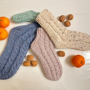 Könnte beinhalten: Vier Paar gestrickte Socken in verschiedenen Farben und Mustern, darunter Blau, Grün, Beige und Rosa. Die Socken liegen auf einer weißen Oberfläche mit einigen Walnüssen, die herumliegen.