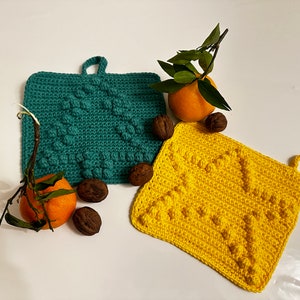 Könnte beinhalten: Zwei gehäkelte Topflappen, einer türkis und einer gelb, mit einem strukturierten, unebenen Design. Die Topflappen haben einen Schlaufe-Griff. Es gibt zwei Orangen und drei Walnüsse auf einer weißen Oberfläche.
