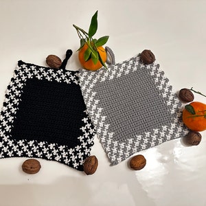 May include: Two crocheted pot holders with a houndstooth pattern border. One is black with a white border and the other is gray with a white border.