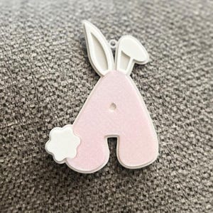 Puede incluir: Una letra A rosa con forma de conejo con orejas blancas y una cola blanca esponjosa. La letra está hecha de un material texturizado.