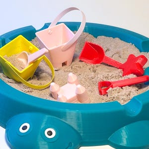 May include: A blue turtle-shaped sandpit filled with sand and colourful toys. The toys include a yellow bucket with a pink scoop, a pink bucket, and red shovels. The sandpit is designed for children's play.