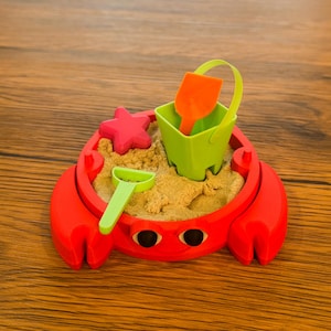 May include: A red crab-shaped sand mold filled with sand, a green bucket with an orange shovel, a pink starfish, and a green shovel. The toy set is on a wooden surface.