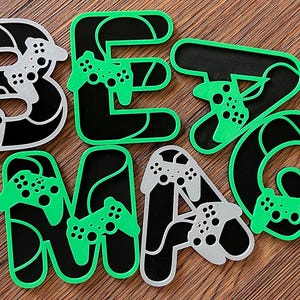 Puede incluir: Letras negras y verdes que deletrean "BE MAG" con un diseño de mando de videojuego. Cada letra tiene un diseño diferente del mando.