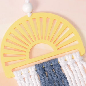 May include: A yellow sun-shaped wooden wall hanging with a white bead at the top. The sun has a cut-out design and is decorated with white and blue yarn tassels.