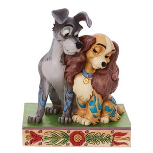 May include: A figurine of two dogs, a gray dog and a tan dog, embracing. The tan dog has brown hair and a blue collar. The gray dog has blue accents. The base is green with floral designs.