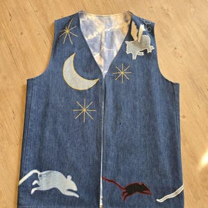 May include: A blue denim vest with embroidered details. The vest features a crescent moon, stars, a flying pig, and two mice.