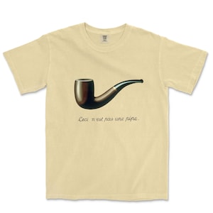 May include: A light yellow t-shirt featuring a dark brown pipe with a black stem. The text "Ceci n'est pas une pipe" is printed below the pipe in a cursive font. The shirt is made of a soft material.