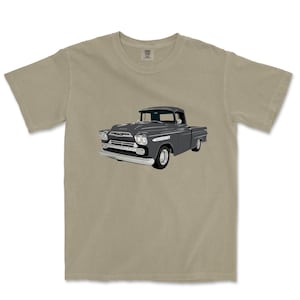 May include: A tan-colored t-shirt featuring a gray illustration of a classic pickup truck. The truck has a detailed design with a white front bumper and chrome accents. The shirt has a crew neck and short sleeves, suitable for casual wear.