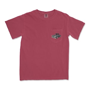 May include: A maroon pocket t-shirt with a small embroidered image of a classic gray pickup truck on the pocket. The shirt has short sleeves and a crew neck. The fabric appears to be a soft, comfortable material.