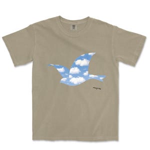 May include: A tan-colored t-shirt featuring a blue bird silhouette filled with a sky and cloud design. The word "magritte" is written in black script on the lower right side of the shirt. The shirt has a crew neck and short sleeves.