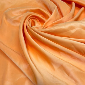 Könnte beinhalten: Eine Nahaufnahme eines pfirsichfarbenen Stoffes mit einer glatten, seidenartigen Textur. Der Stoff ist drapiert und gefaltet und erzeugt weiche, fließende Linien.