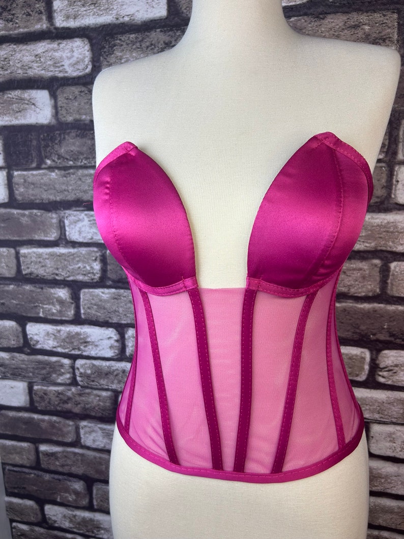 May include: A pink satin and mesh corset with a plunging neckline and boning. The corset has a satin top and a mesh bottom with satin trim.
