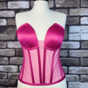 May include: A pink satin and mesh corset with a sweetheart neckline. The corset has boning and a hook and eye closure in the back.