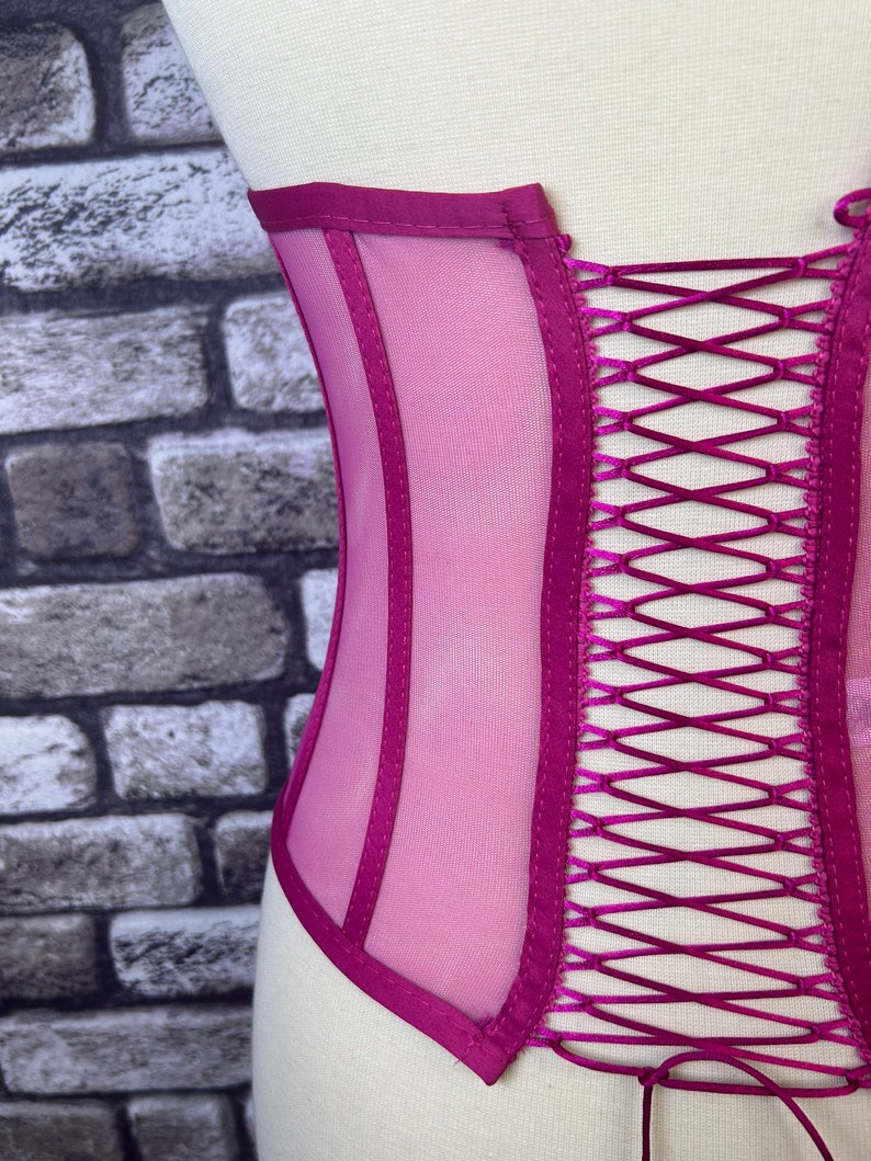 May include: A magenta mesh corset with satin ribbon lacing in the back. The corset has a satin trim and is designed to cinch the waist.