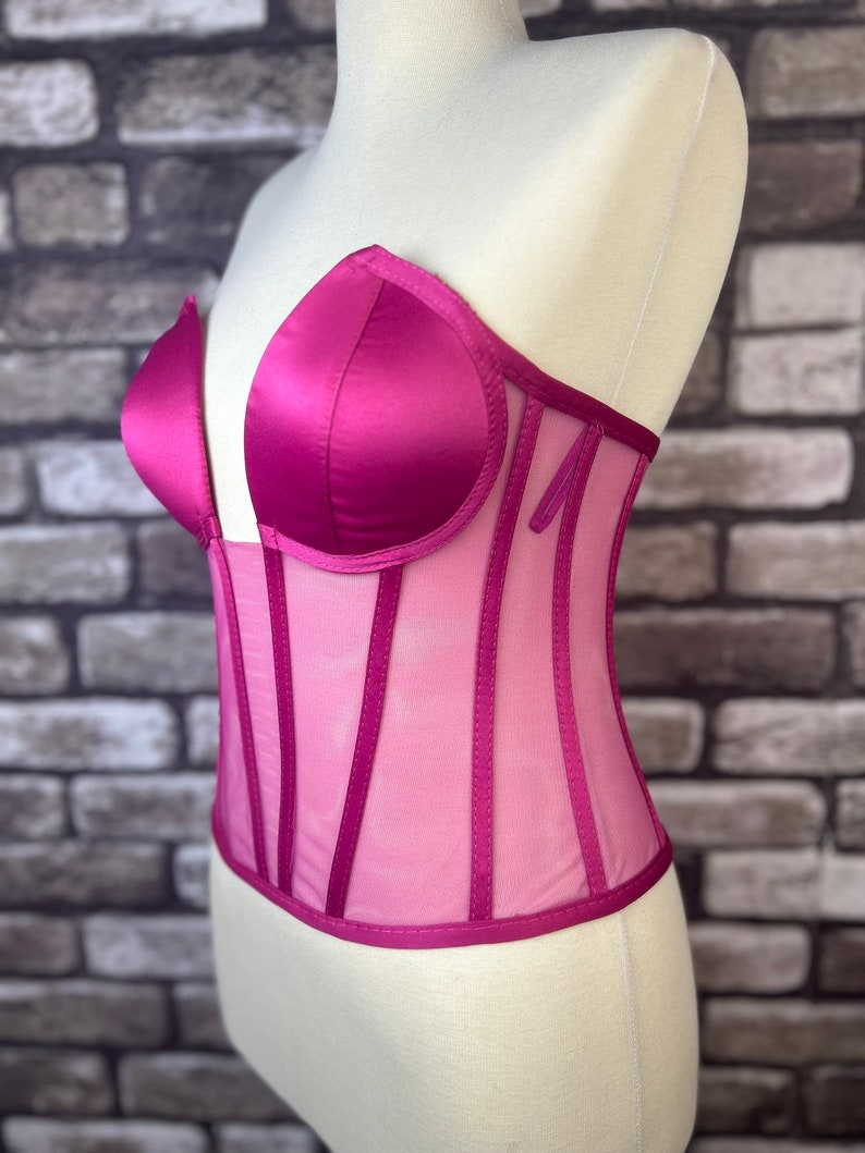 May include: A pink satin and mesh corset with a zipper closure. The corset has a sweetheart neckline and a boned bodice.