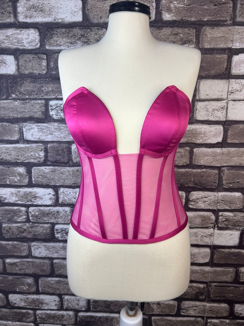 May include: A pink satin and mesh corset with a sweetheart neckline. The corset has boning and a hook and eye closure in the back.
