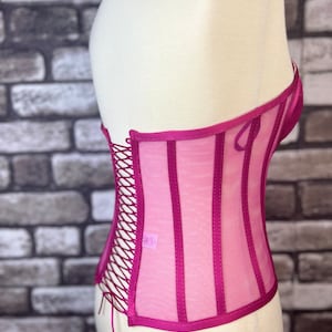 May include: A pink mesh corset with a lace-up back and satin trim. The corset has a boned structure and is designed to cinch the waist.
