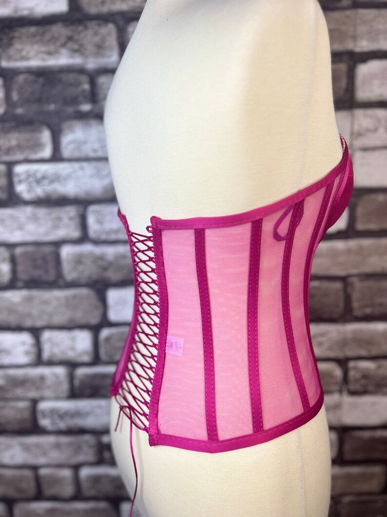 May include: A pink mesh corset with a lace-up back and satin trim. The corset has a boned structure and is designed to cinch the waist.