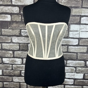 Puede incluir: Un corsé blanco de malla con ribete de satén. El corsé tiene un diseño sin tirantes y una estructura con varillas.