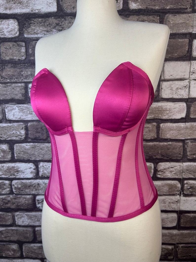 May include: A pink satin and mesh corset with a plunging neckline and boning. The corset has a sweetheart neckline and is strapless.