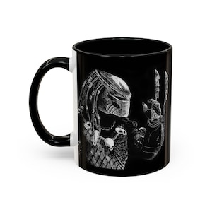 May include: Black ceramic coffee mug with a white illustration of a Predator character with a skull and bones on its chest. The Predator is making a hand gesture with its right hand.