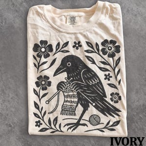 May include: An ivory t-shirt featuring a black graphic print of a crow knitting with two needles and a ball of yarn. The design is framed by black floral motifs and leaves. The shirt has a crew neckline and a Comfort Colors brand label at the neck.