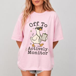 May include: A light pink t-shirt with the text "Off To Actively Monitor" and a cartoon goose wearing sunglasses and holding a clipboard. The goose has a name tag that says "Teacher in Charge".