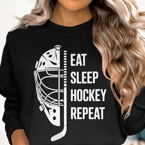 Puede incluir: Sudadera negra con un gráfico blanco de máscara de portero de hockey y palo. El texto "EAT SLEEP HOCKEY REPEAT" está impreso en blanco. Una camiseta informal para los aficionados al hockey.