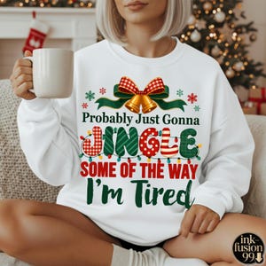 Puede incluir: Sudadera blanca con un diseño navideño. El diseño incluye un lazo a cuadros rojo y verde, campanas doradas, copos de nieve y el texto "Probably Just Gonna Jingle Some Of The Way I'm Tired."