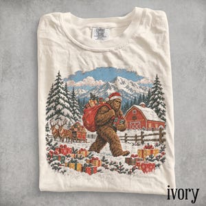 May include: An ivory t-shirt featuring a graphic of a brown Bigfoot wearing a Santa hat, carrying a red sack of gifts and a wrapped present. The scene includes a snowy landscape, pine trees, a red barn, and reindeer pulling a sleigh.